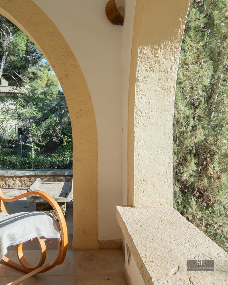 A classic bentwood rocking chair sits on a stone arched terrace overlooking a lush green garden.