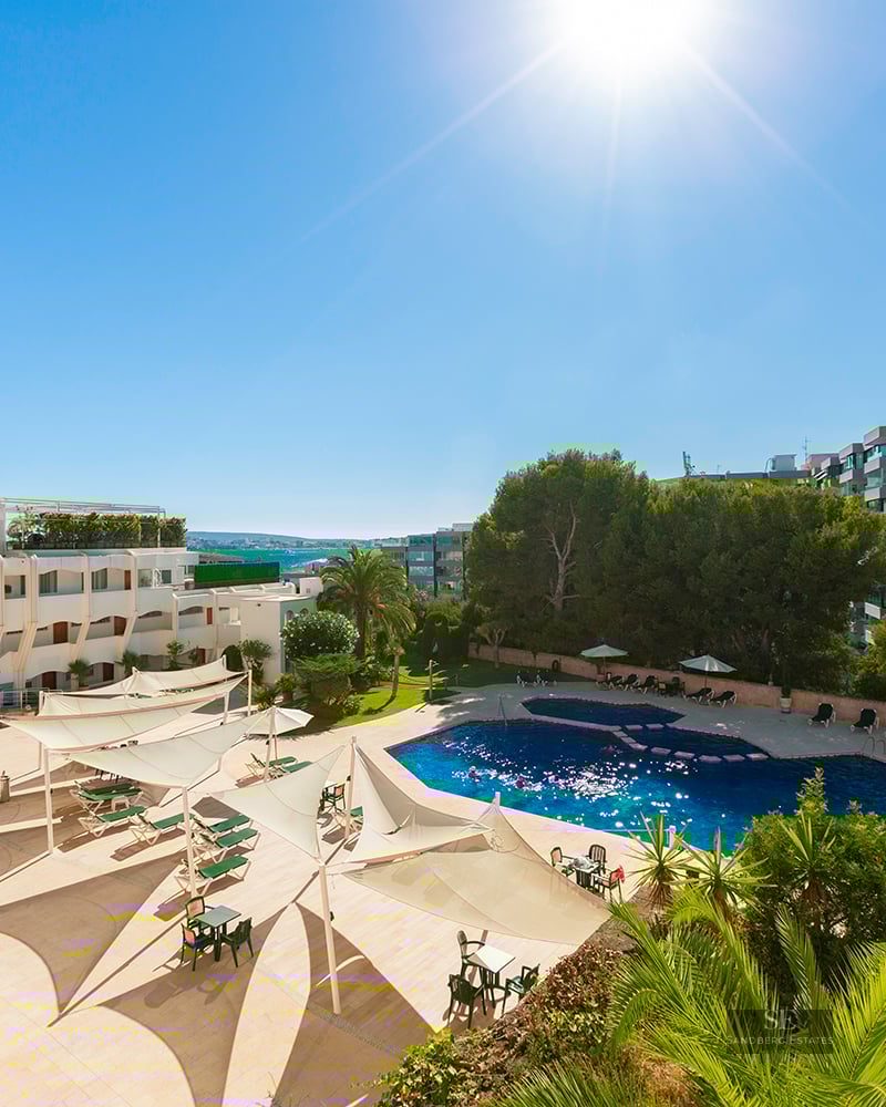 Large blue swimming pool surrounded by beige terrace, white sun sails, palm trees, and resort buildings under a bright sun.
