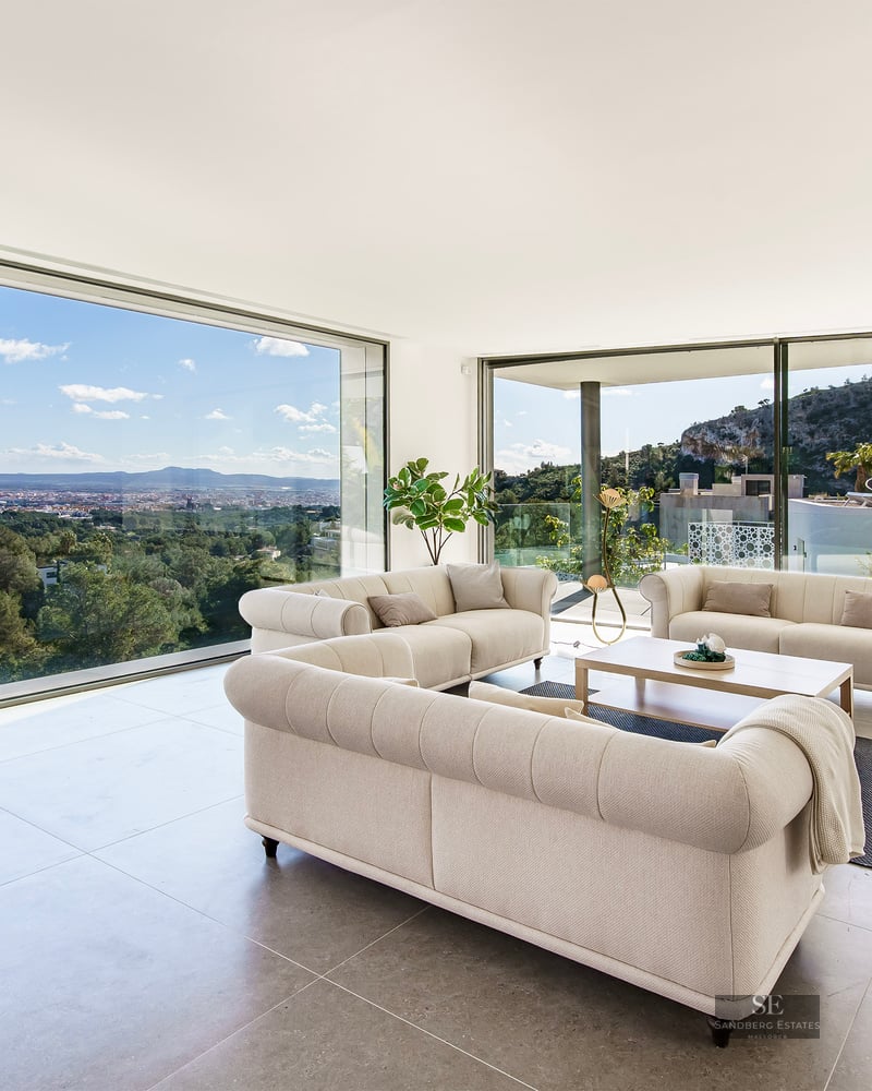 Modern living room with three beige tufted sofas and floor-to-ceiling windows overlooking a city valley and mountains.