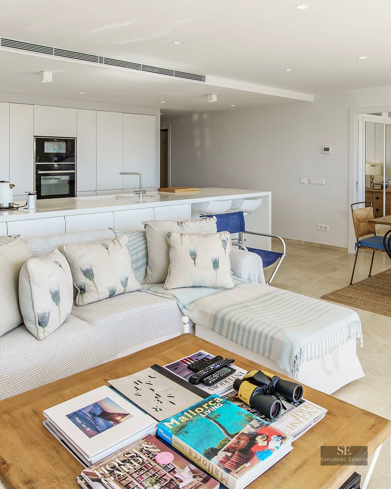 Bright open-plan living room with white sofa, wooden coffee table, dining set, and minimalist kitchen in the background.