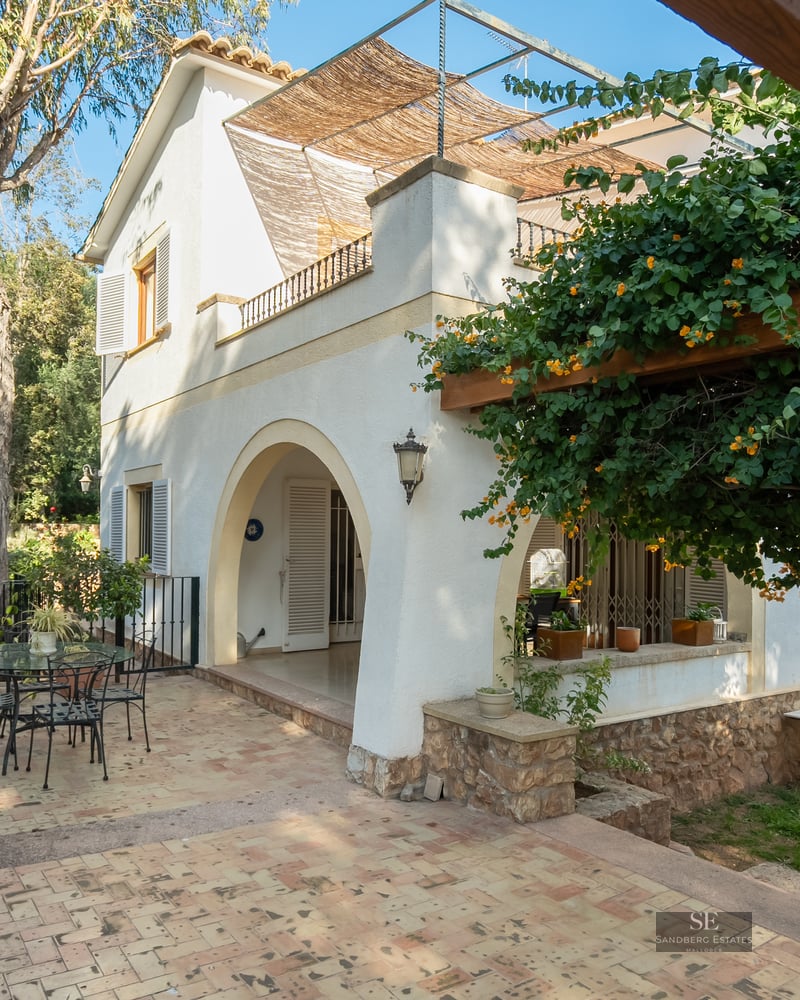 Sunny tiled patio with a flowering wooden pergola and white villa facade featuring arched entrance.