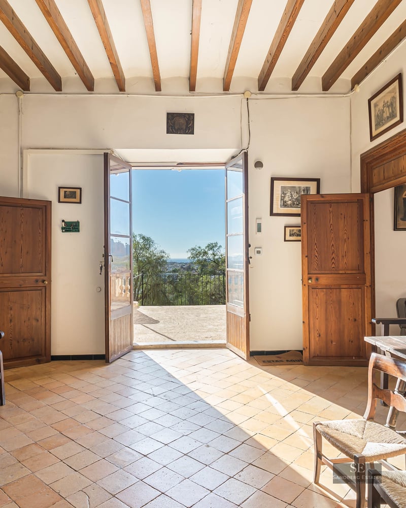 Traditional hallway with wooden beams, terracotta floors, and open doors leading to a sunny terrace.