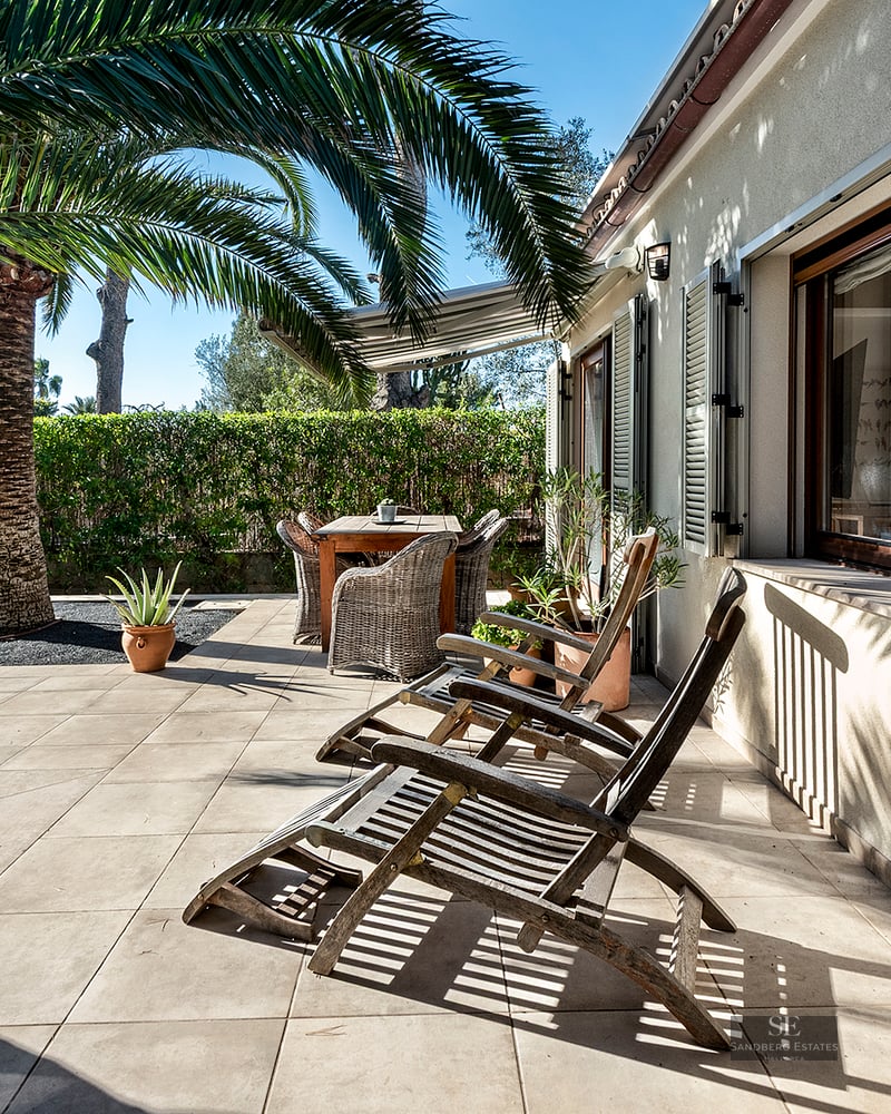 Sunny tiled patio with wooden lounge chairs, a dining set, and a large palm tree against a beige house wall with shutters.