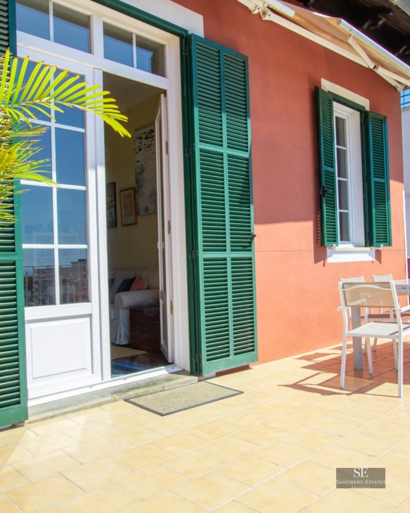 A sunny rooftop terrace featuring terracotta walls, green shutters, white dining furniture, and lush potted plants.