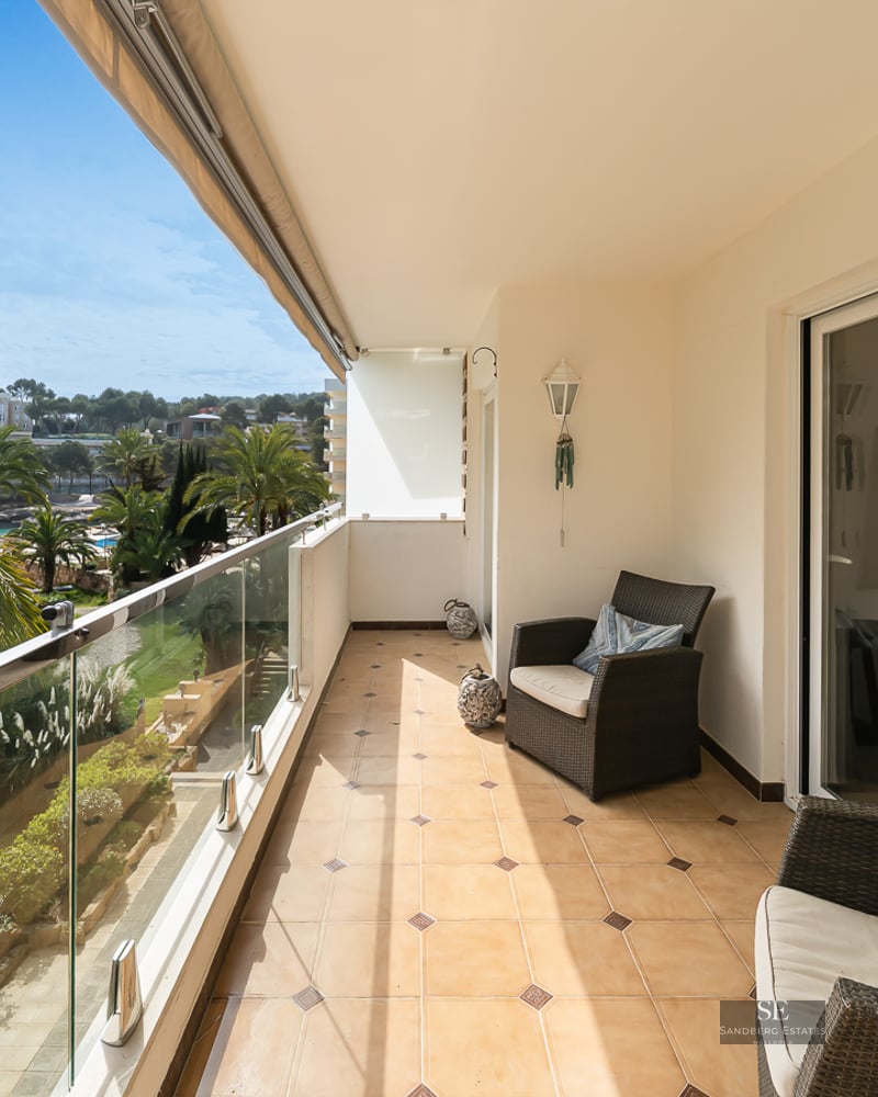 Sunlit balcony with wicker chairs, glass railing, and a scenic view of lush palm trees and a garden.