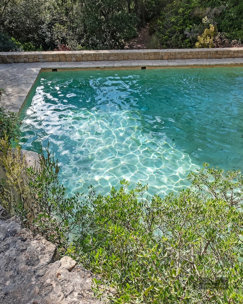 Elevated view of a turquoise stone pool surrounded by dense green forest and a stone terrace with a sun umbrella.