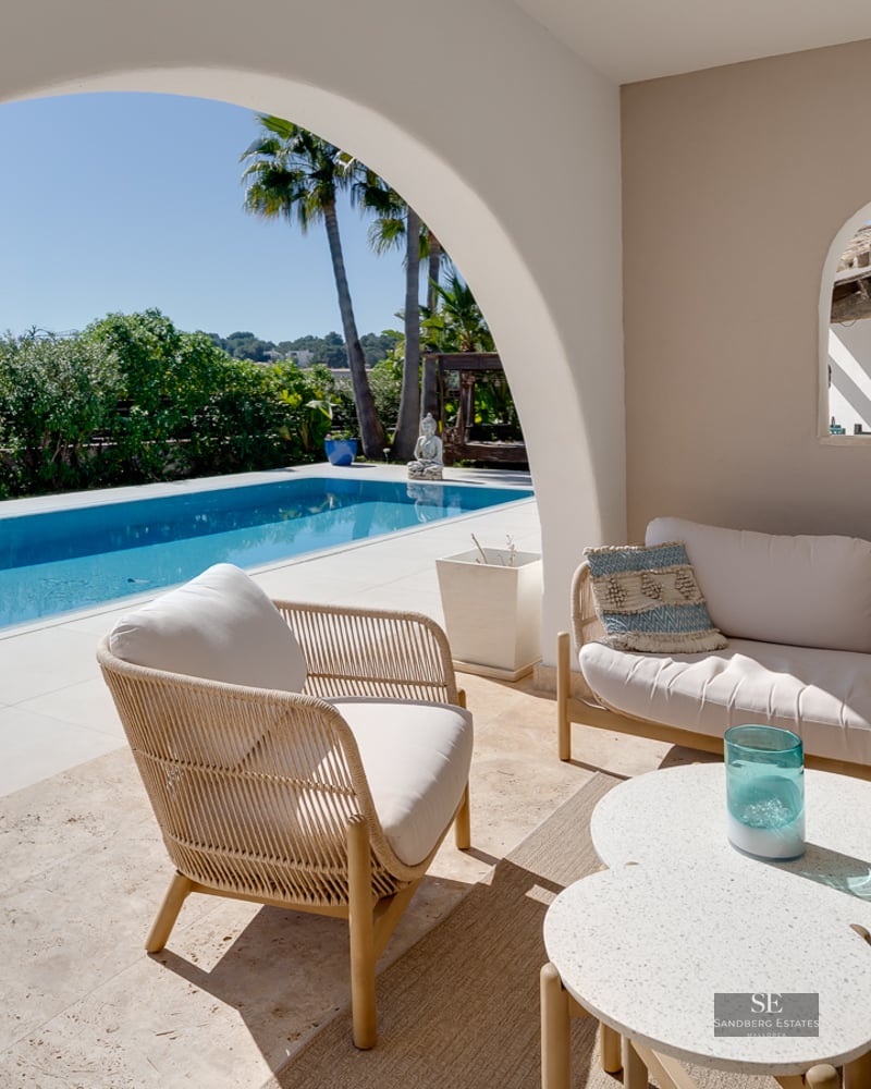 A covered terrace with white lounge furniture looking out through a large archway to a blue swimming pool.