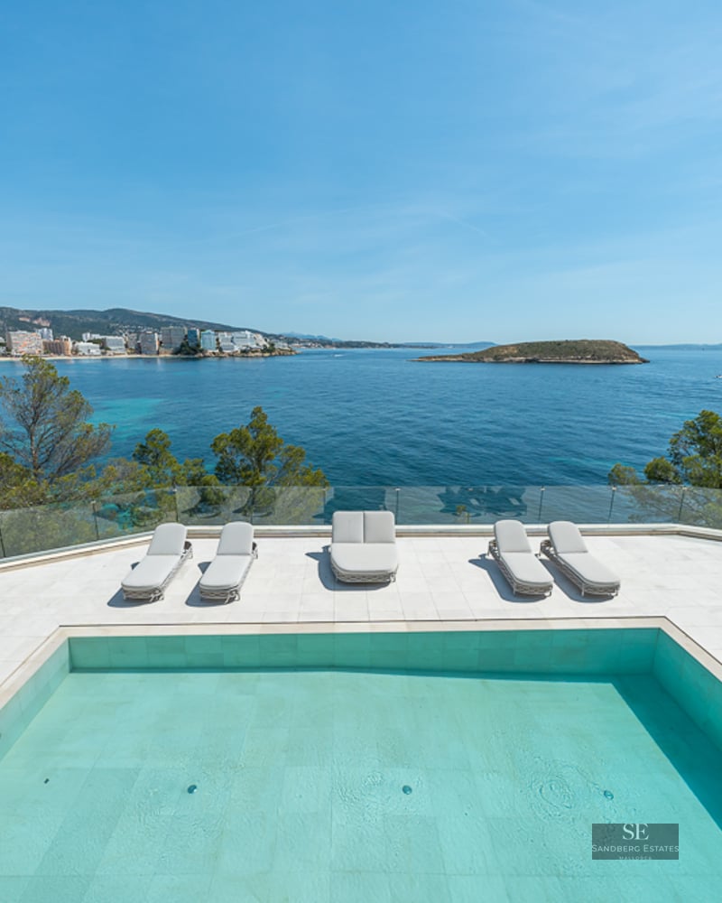 Modern swimming pool and sun loungers on a white terrace overlooking a turquoise bay and distant coastline.