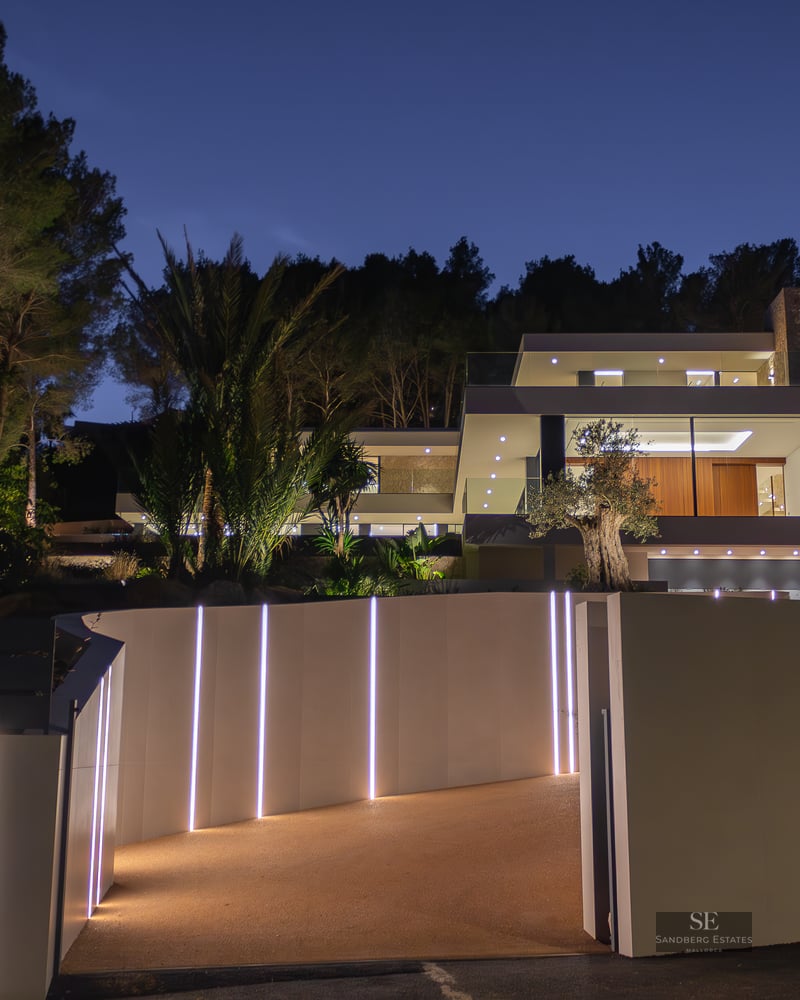 Modern white luxury villa at night with illuminated LED driveway, glass walls, and lush palm trees against a dark sky.