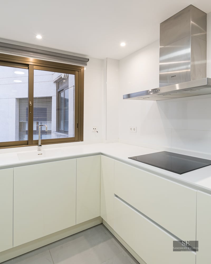 Minimalist white kitchen featuring L-shaped counter, induction stove, stainless steel extractor, and window.