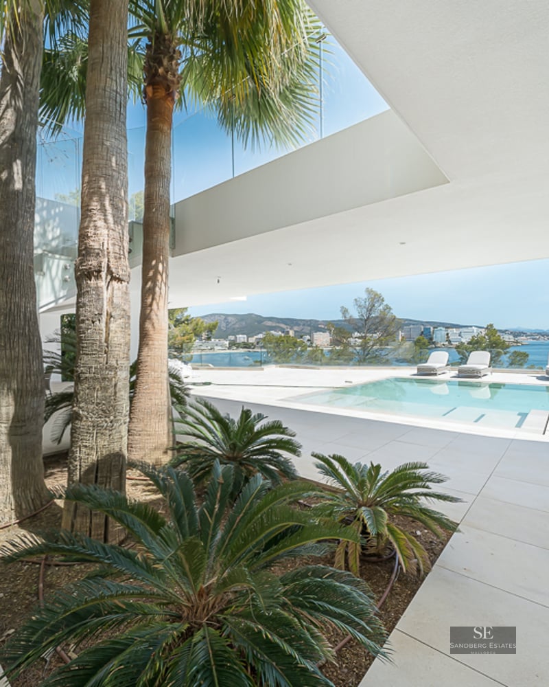 Modern villa terrace featuring a pool, palm trees, and glass railings overlooking the blue Mediterranean sea.