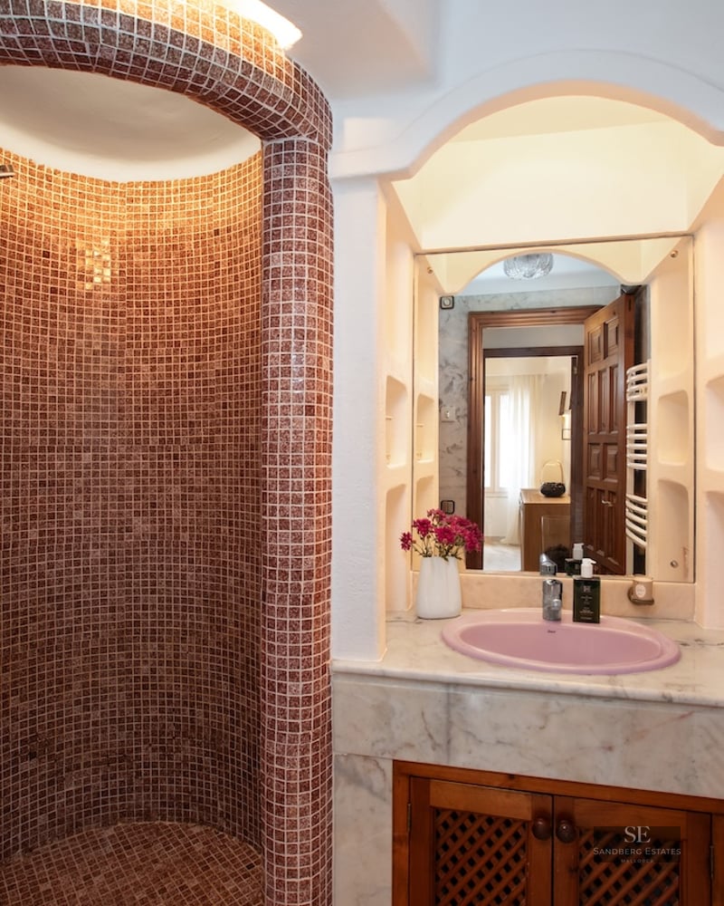 A curved walk-in shower with reddish mosaic tiles next to a marble vanity with a pink sink and a mirror.