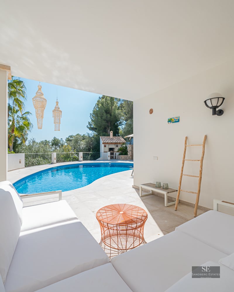 Une élégante terrasse couverte avec des canapés blancs donnant sur une piscine courbe entourée de verdure.
