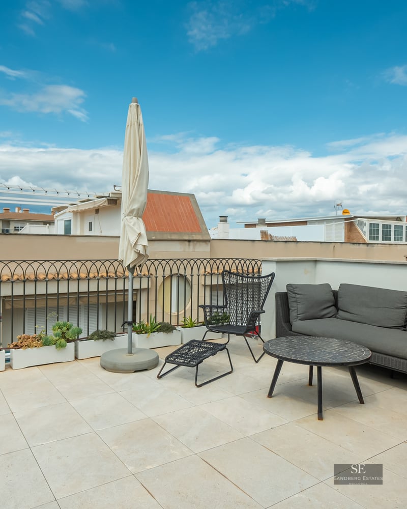Rooftop terrace with grey sofa, black chairs, and a white umbrella under a blue sky.