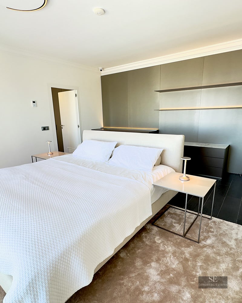 Modern bedroom featuring a large white bed, minimalist nightstands, a taupe feature wall, and a plush beige rug.