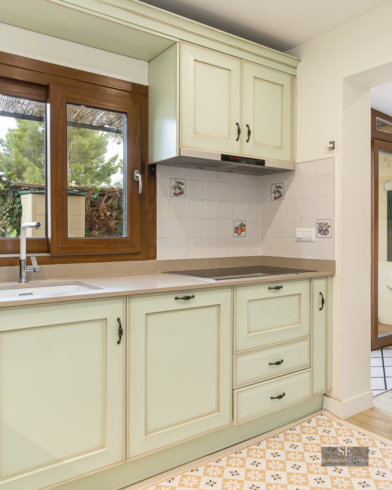 Bright kitchen with mint green cabinets, wooden window, and patterned floor leading to a dining area with wicker chairs.