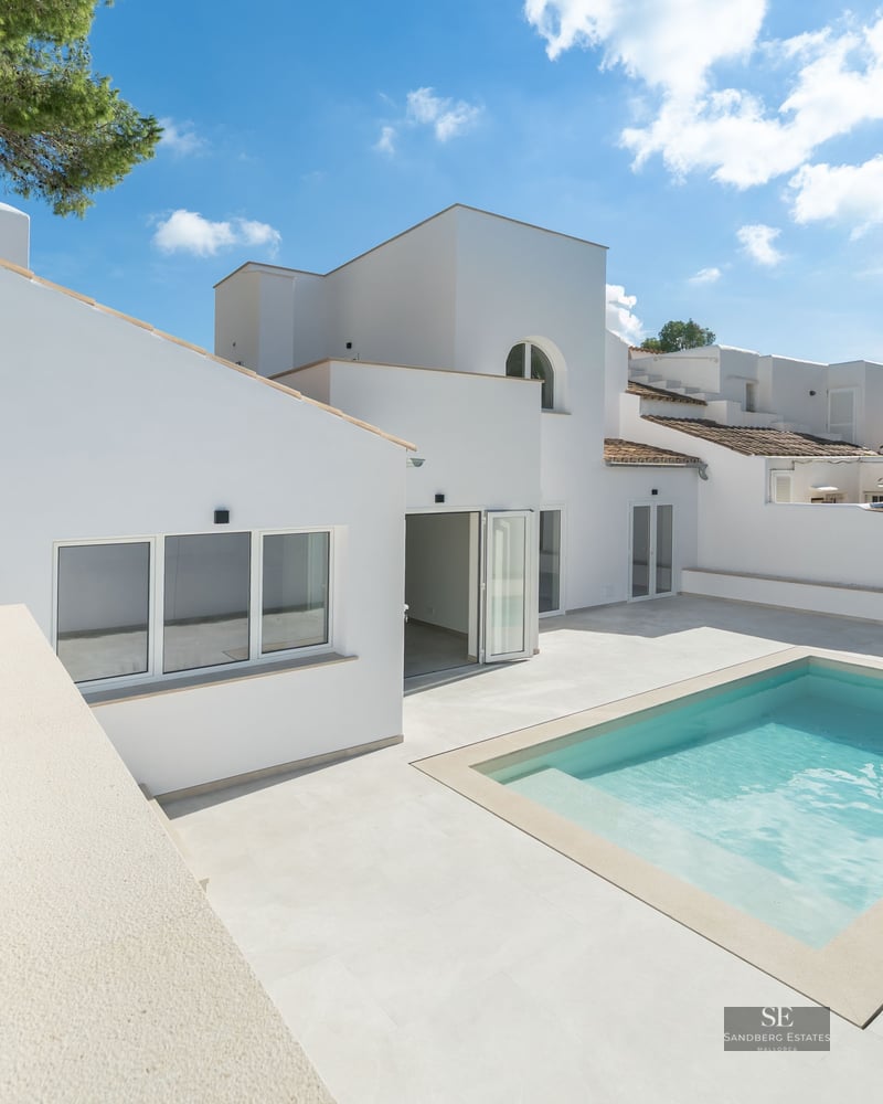 Modern white Mediterranean villa featuring a bright swimming pool, spacious stone patio, and outdoor kitchen area.