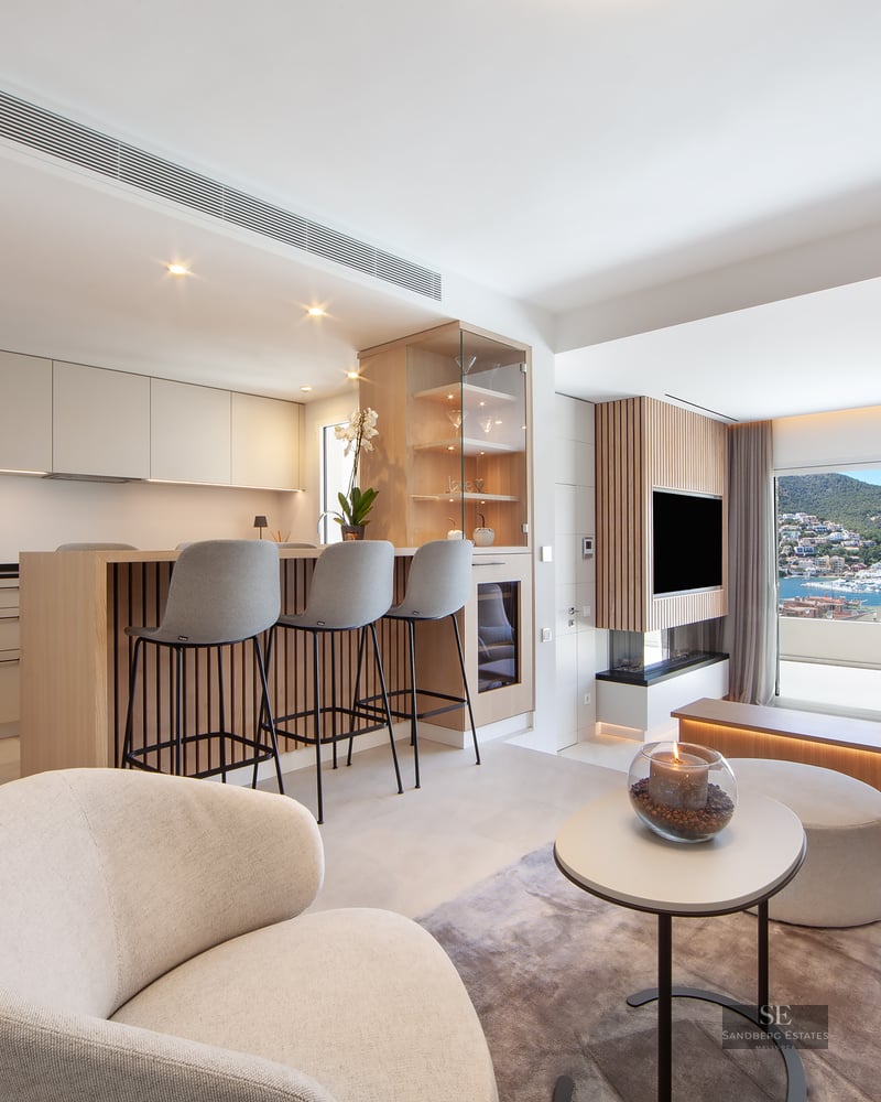 A bright, modern open-plan living room and kitchen featuring beige cabinetry, an island with stools, and a large window with a marina view.