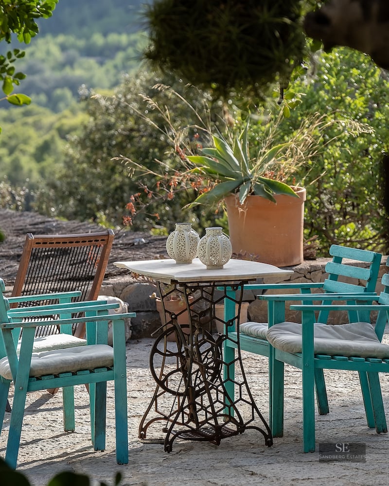 Vintage marble table and teal wooden chairs on a stone patio surrounded by lush Mediterranean greenery and mountain views.