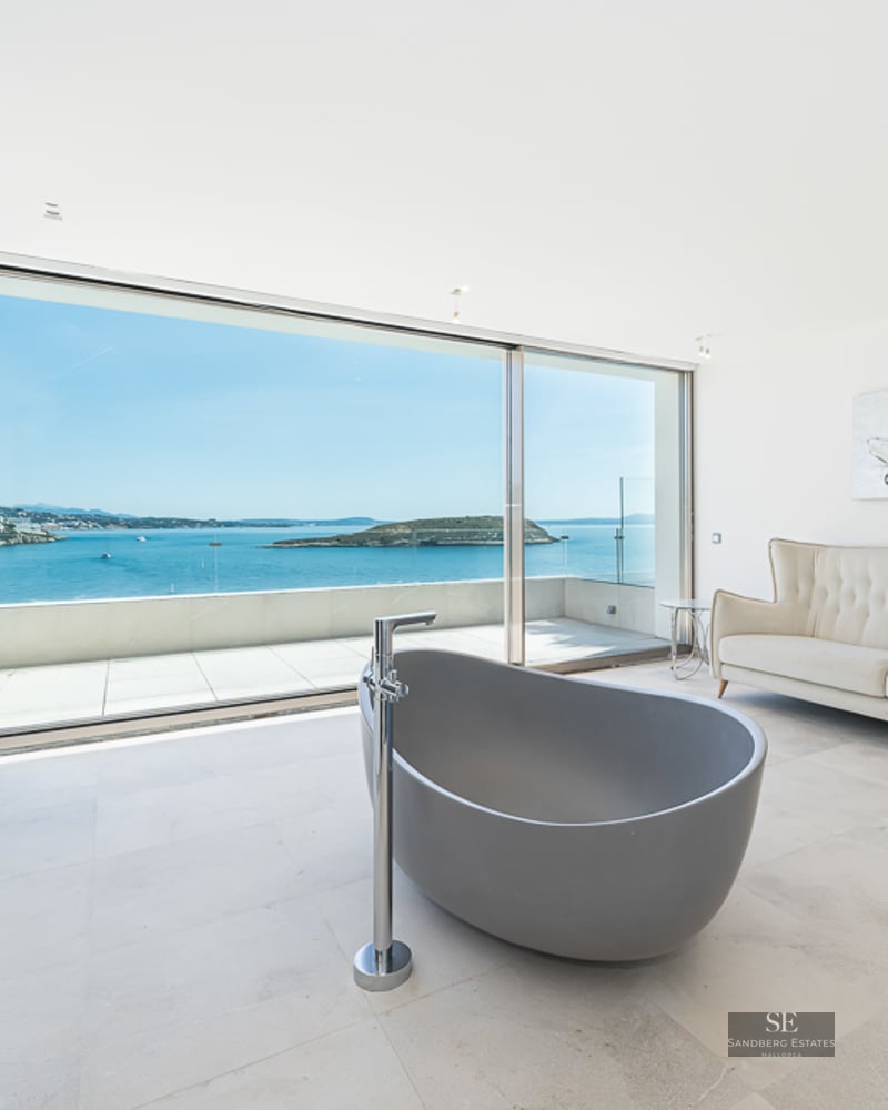 Modern master bathroom featuring a freestanding grey tub, cream sofa, and floor-to-ceiling windows overlooking the ocean.