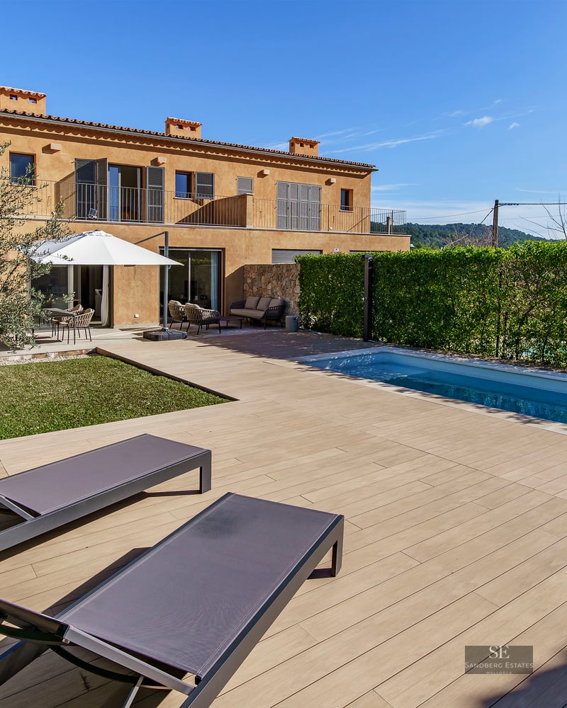 Modern outdoor pool and wooden deck with sun loungers next to a terracotta villa under a clear blue sky.