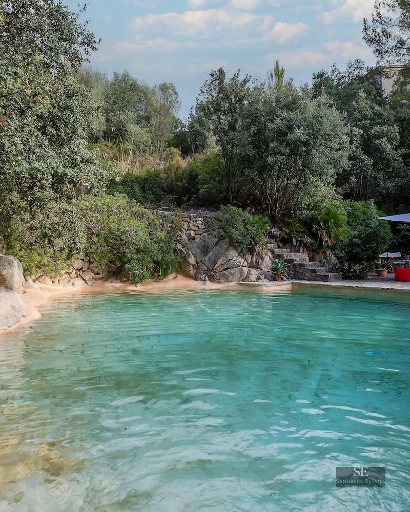 A lagoon-style pool with turquoise water, natural rock edges, and a lounge area with an umbrella and red accents.
