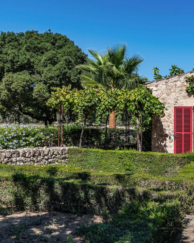 Manicured hedges and stone walls leading to a rustic stone house with red shutters and lush greenery under a blue sky.