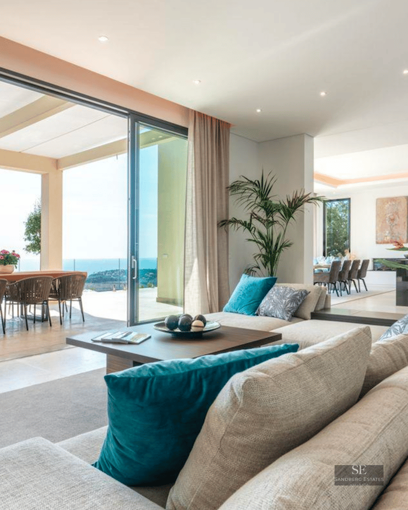 Spacious modern living room with large beige sofa, floor-to-ceiling glass doors opening to a terrace with sea views.