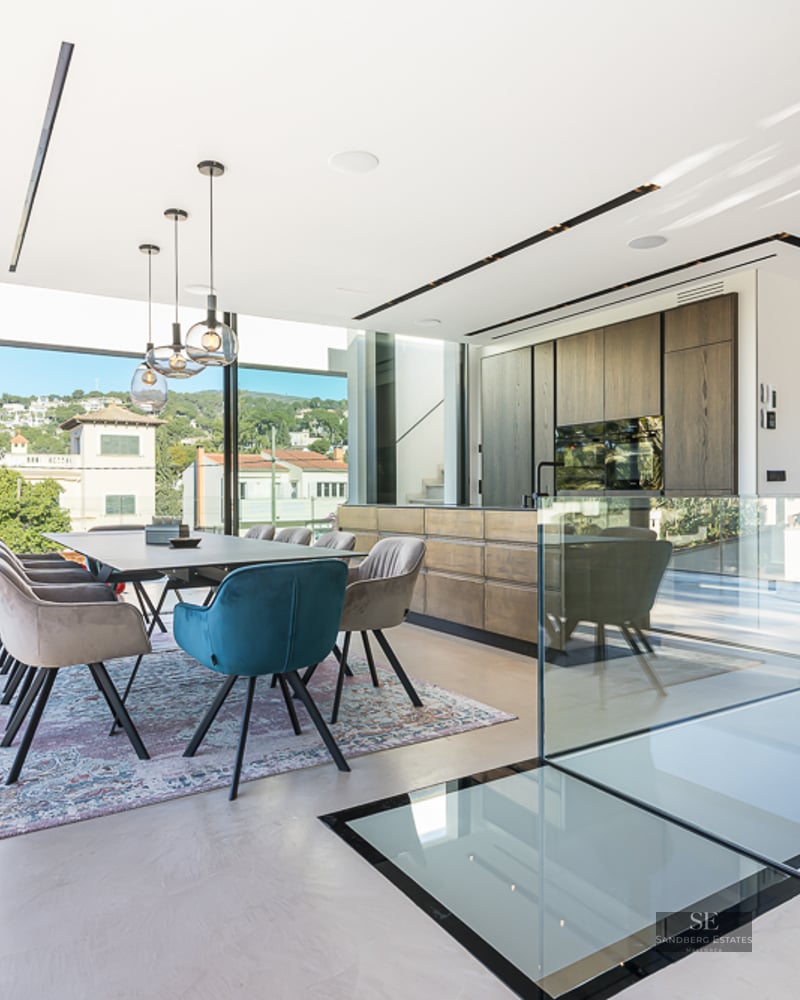 Modern dining room with large windows overlooking scenery, featuring a long table, velvet chairs, and open kitchen.
