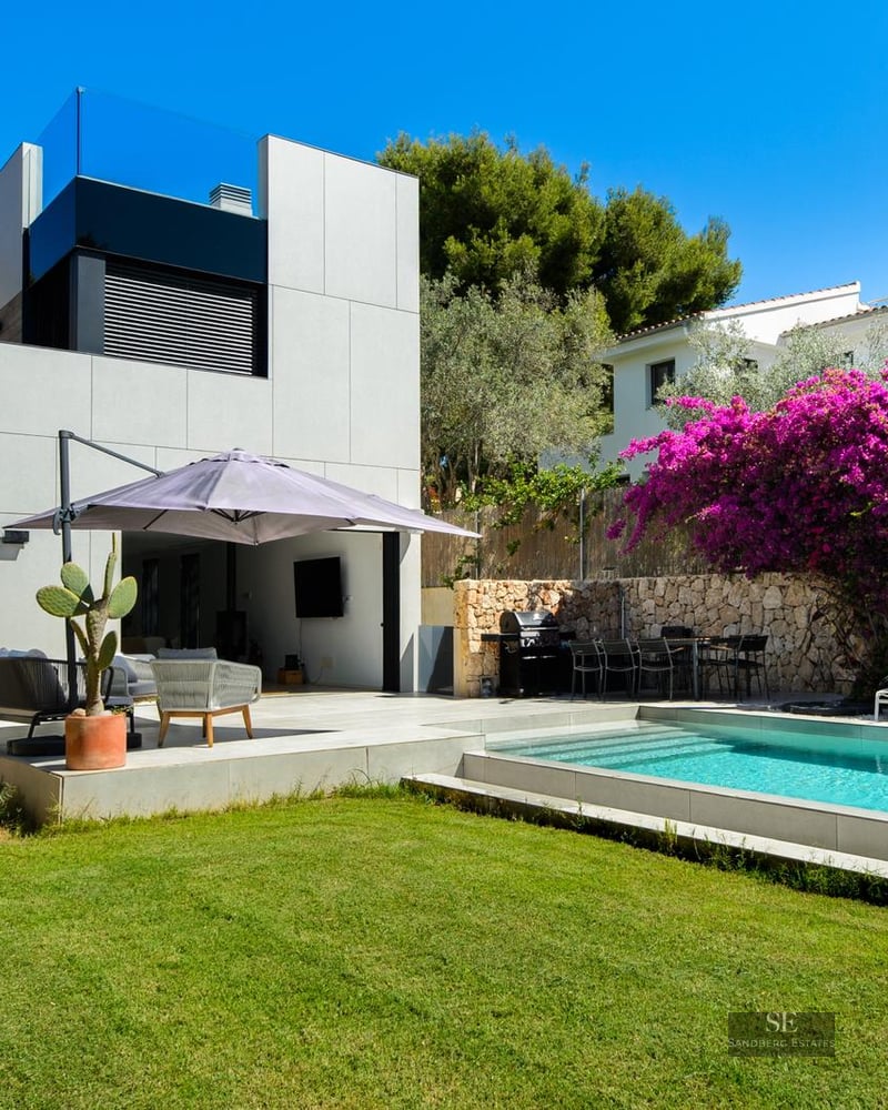 Modern villa exterior with a blue rectangular pool, green lawn, and bright purple bougainvillea against a stone wall.