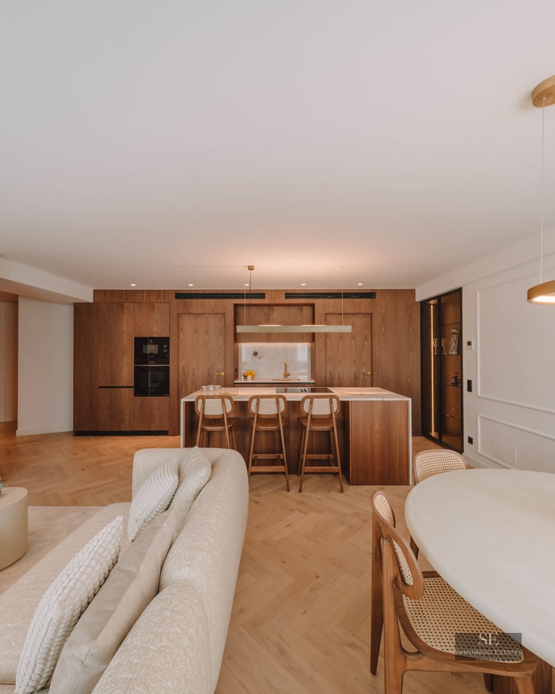 Spacious luxury living room featuring a beige sofa, herringbone wood floors, and an integrated natural wood kitchen.