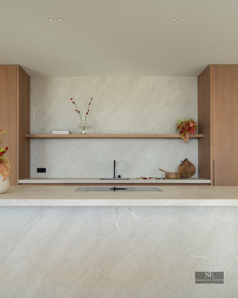 Modern kitchen featuring a large beige stone island, natural wood cabinets, and a matching stone backsplash.