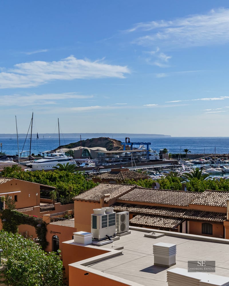 Elevated view of a Mediterranean marina with terracotta roofs, white yachts, and the blue sea under a clear sky.