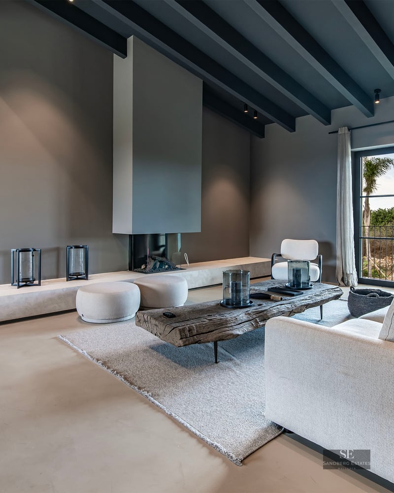 Spacious modern living room with grey ceiling beams, beige sofa, rustic wood table, and built-in minimalist fireplace.