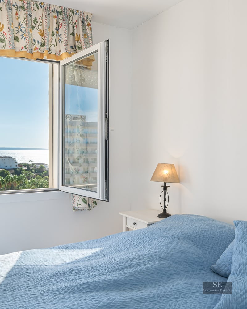 A sunlit bedroom featuring a blue quilted bed and an open window looking out over trees and the sparkling sea.