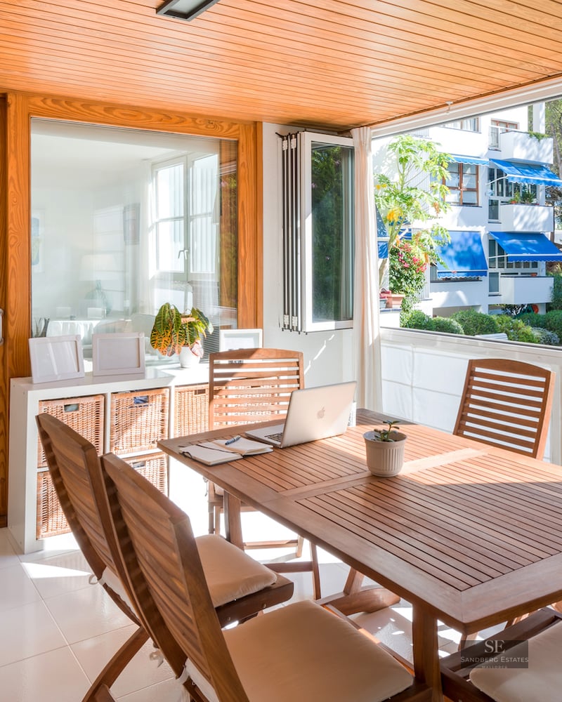 Bright enclosed wooden terrace with dining table and laptop, overlooking a pool and garden through large windows.