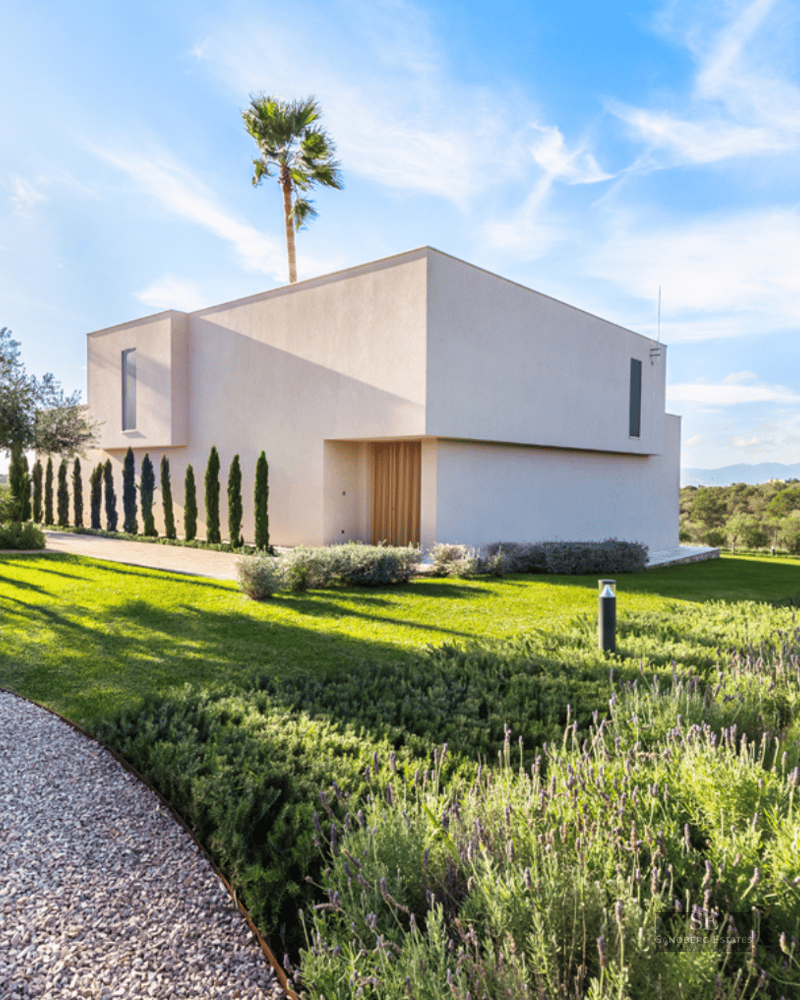 Exterior of a white minimalist villa featuring lavender gardens, green lawn, and palm trees under a bright blue sky.