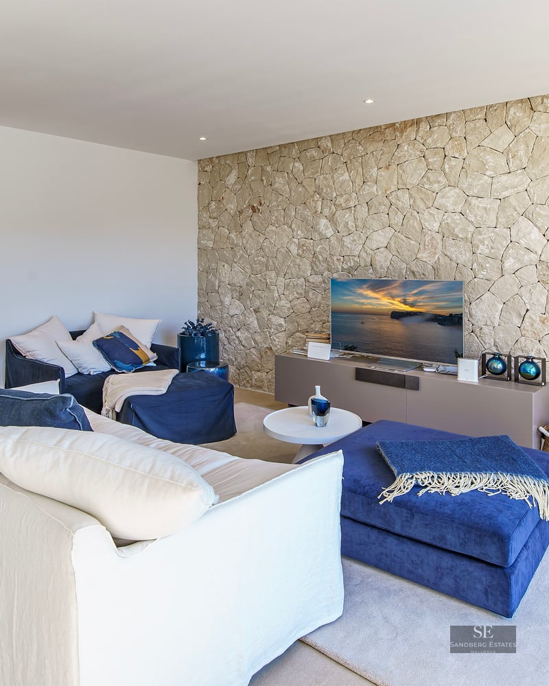 Contemporary living room featuring a white sofa, navy blue ottomans, and a textured natural stone wall with a TV.