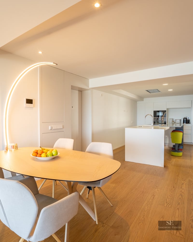Minimalist dining area with a light wood table, grey chairs, and a striking arched floor lamp, leading to a modern kitchen.