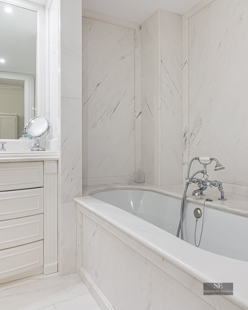 Bright master bathroom featuring white marble walls, a built-in tub with chrome faucets, and a white vanity with drawers.