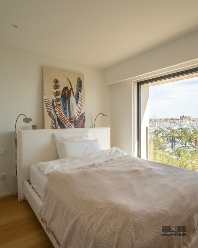 Modern bedroom with a white bed and a large window overlooking a sunny marina with palm trees.
