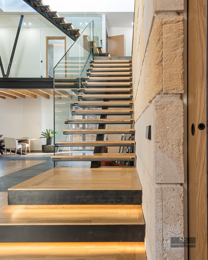 Modern open-plan living room featuring a wooden floating staircase with LED lights, glass railings, and a mezzanine.