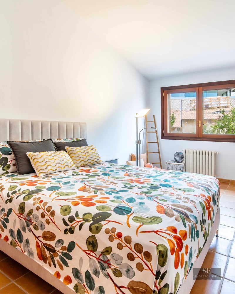A bedroom featuring floral bedding, a grey upholstered headboard, terracotta tile floors, and a wooden window.