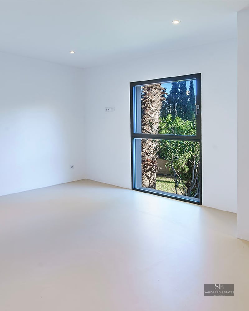 Bright, empty minimalist room with white walls, light flooring, and a vertical window framing a palm tree outside.