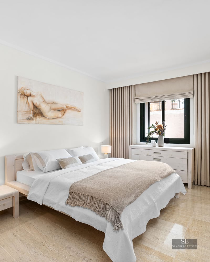 Chambre lumineuse avec lit en bois clair, sol en marbre, murs blancs et un grand tableau au-dessus du lit.