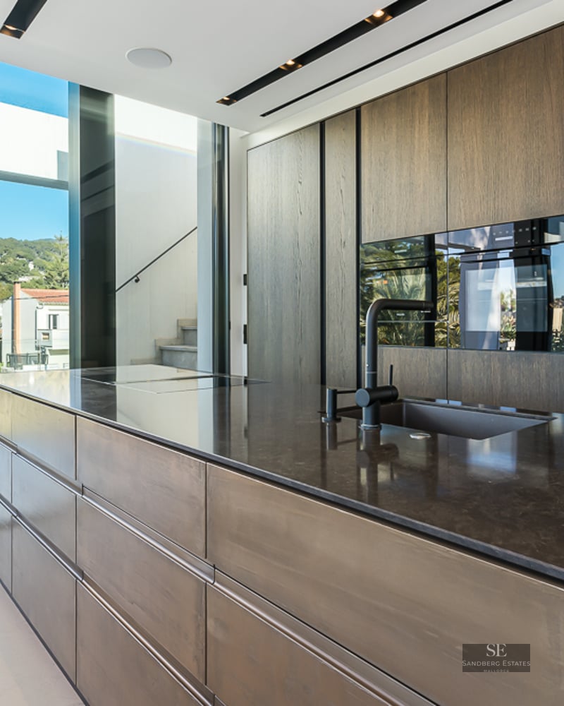Modern kitchen featuring a large dark stone island, wood cabinetry, and floor-to-ceiling windows with a landscape view.