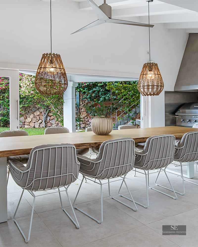 Large live-edge wooden dining table with designer chairs and woven pendant lights next to a stone-walled outdoor kitchen.