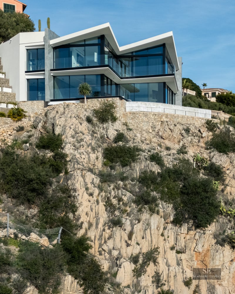 Modern white villa with large glass walls built on a steep rocky cliffside under a clear blue sky.
