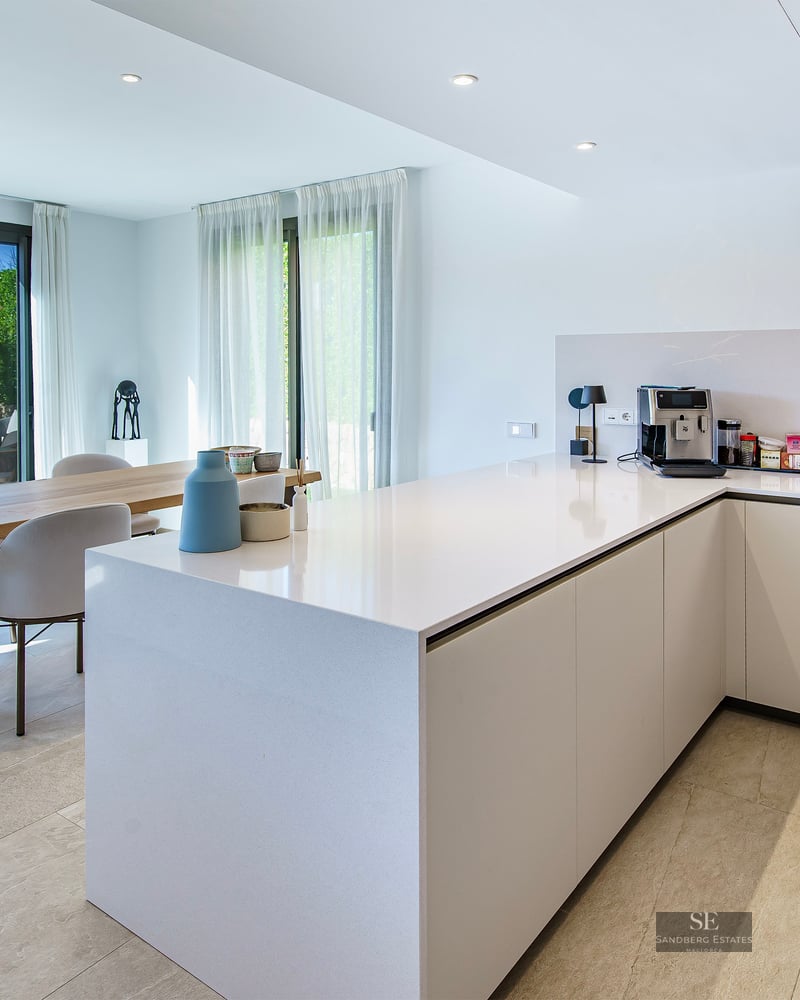 Minimalist white kitchen featuring a large island, wooden dining table, and glass sliding doors leading to a sunny garden.