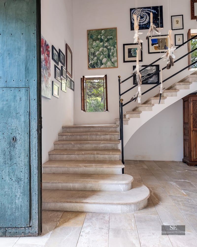 Entrance hall with a large distressed blue door, stone stairs leading up, and a gallery of paintings on white walls.